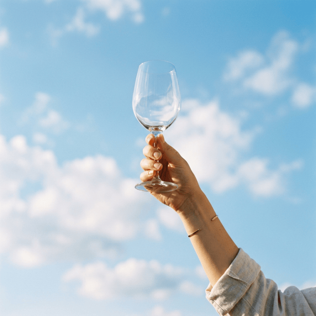 Hand holding an empty wine glass against a blue cloudy sky
